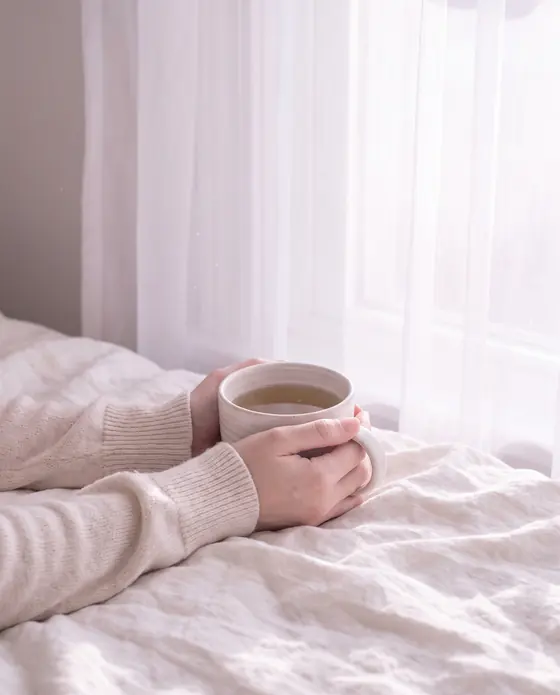 Hände halten eine helle Keramiktasse am Fenster im Morgenlicht